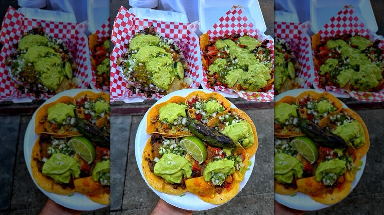 Hand holding plate of tacos with lime wedge and pepper, styrofoam takeout containers of tacos behind