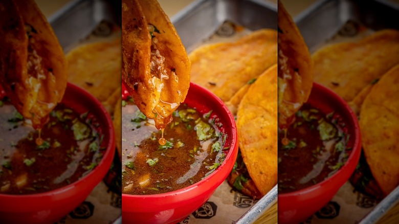 Dipping birria taco in consomme, tacos on tray beside