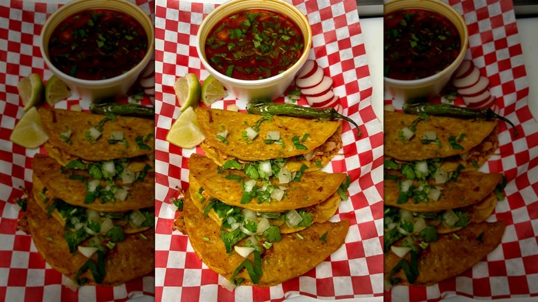 Three quesabirria tacos on red and white checked paper below cup of consommé