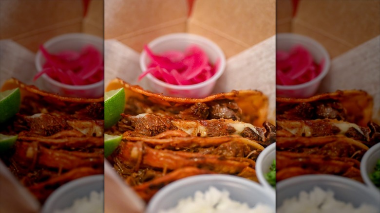 Close-up of birria tacos in takeout box, cup of pickled onions behind