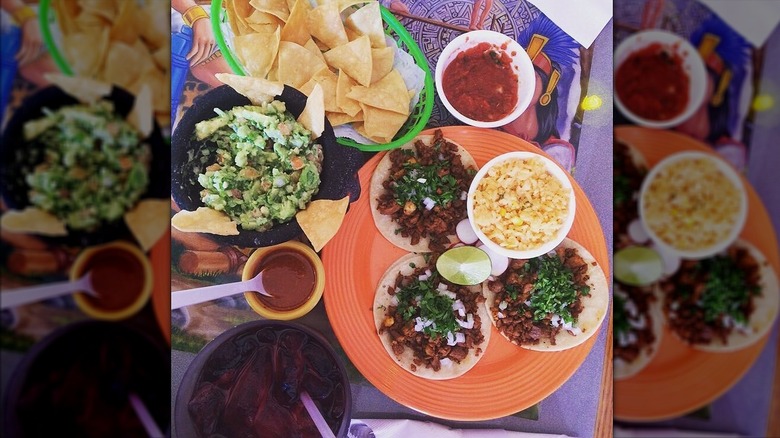 Top-down shot of basket of tortilla chips, bowl of guacamole, plate with three soft tacos and bowl of rice, salsa around