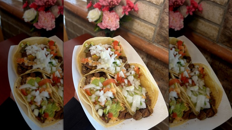 Three soft tacos with meat, tomato, guacamole, and onions held in a small paper basket