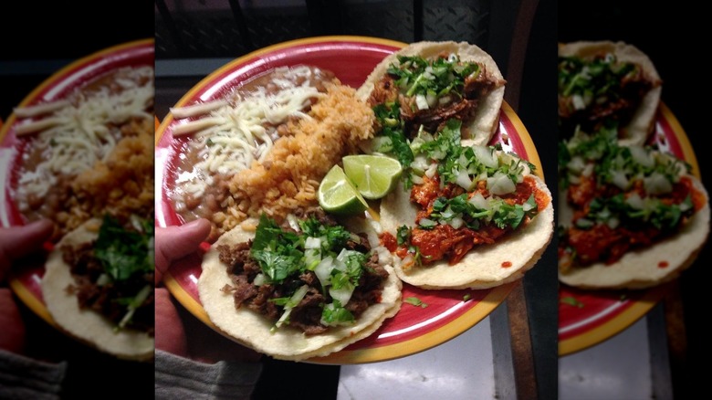 A hand holding a red plate with three soft tacos, refried beans, rice, lime wedges