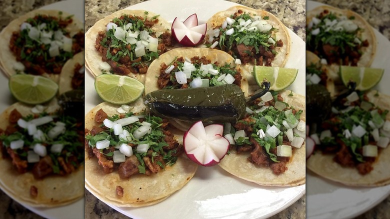 Five soft tacos on a white plate with lime wedges, radish, and a roasted jalapeño