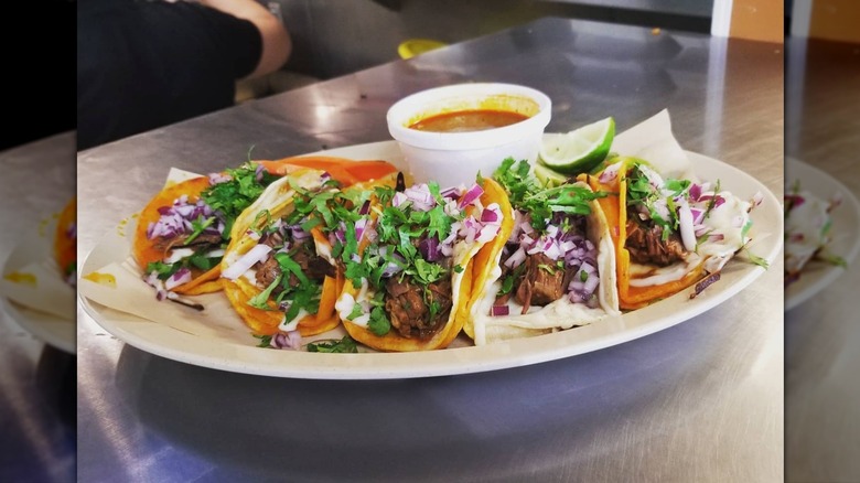 Five soft tacos on a plate next to a styrofoam cup of consommé