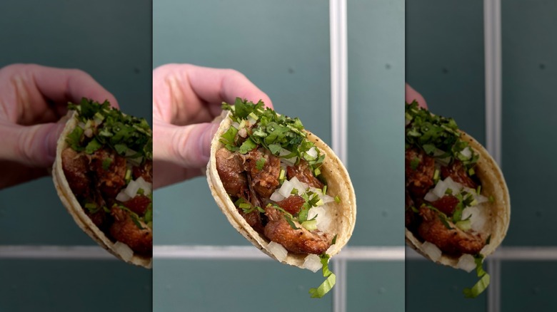 A hand holding a soft taco with meat, onion, cilantro in front of a green wall