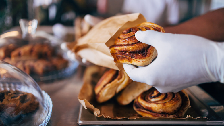 Gloved hand placing cinnamon roll in paper take-out bag
