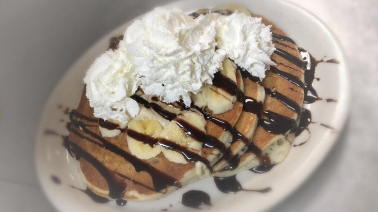 Stack of pancakes with banana slices, chocolate drizzle, and whipped cream on white plate