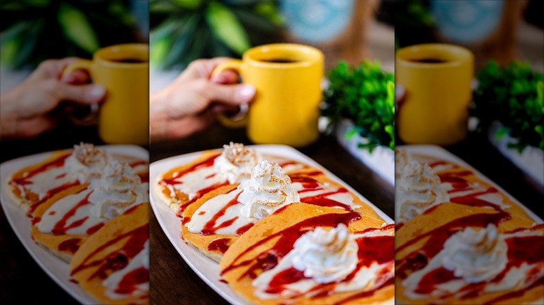 Three pancakes with red drizzle and whipped cream in front of yellow mug