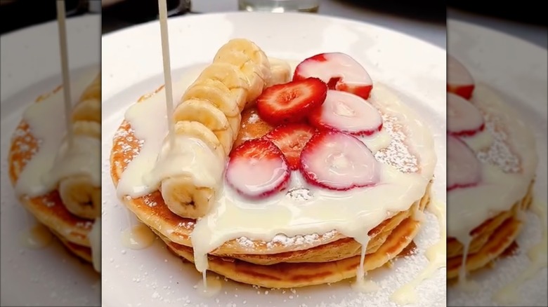 Pouring cream onto tres leches pancakes with banana and strawberry slices on white plate