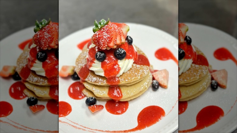 Stack of strawberry pancakes with blueberries and whipped cream on white plate