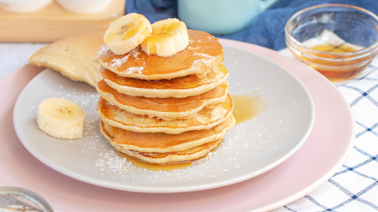 Six pancakes stacked and topped with banana slices and syrup on white plate