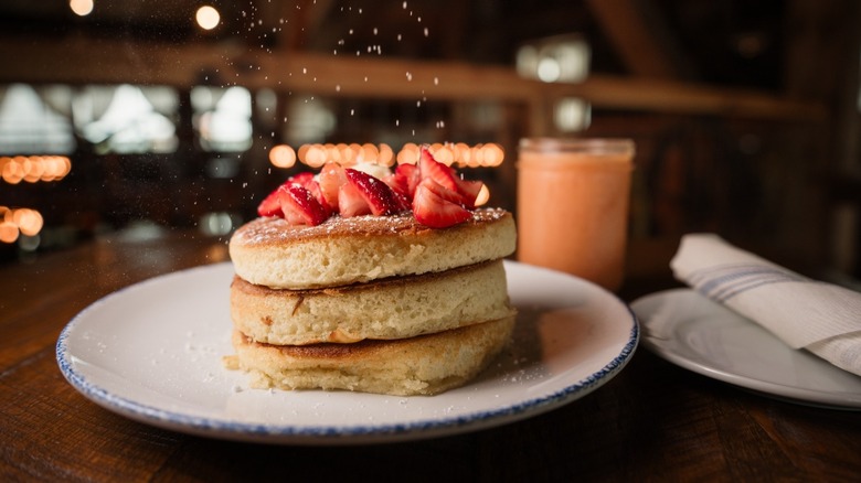 Sprinkling powdered sugar on thick griddle cakes topped with strawberries on white plate