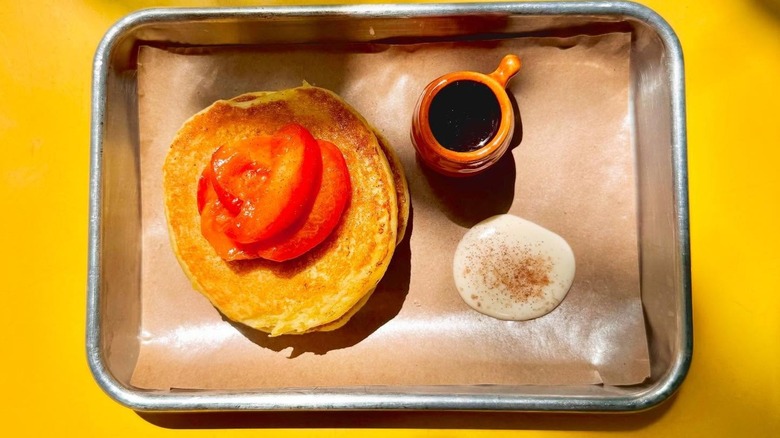 Masa pancakes with peaches next to cream and syrup on metal tray, yellow background
