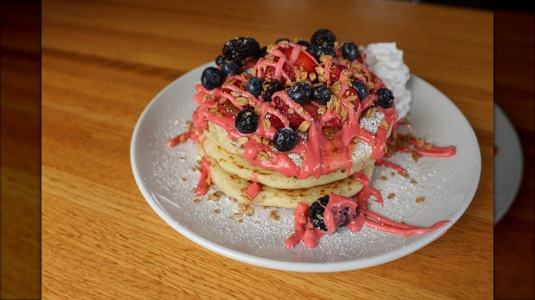 Stack of pancakes with strawberries, blueberries, pink drizzle, nuts on white plate