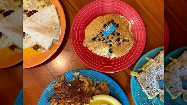 Blueberry pancake with wrapped pat of butter on red plate, surrounded by other plates with breakfast dishes