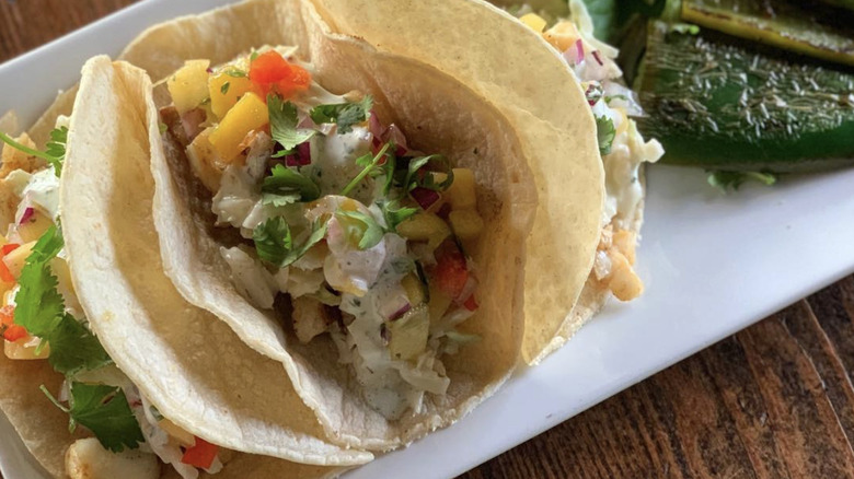 Three fish tacos with pineapple on corn tortillas with peppers on the side