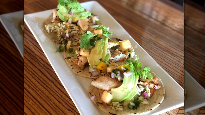 Three fish tacos with avocado on a white rectangular plate