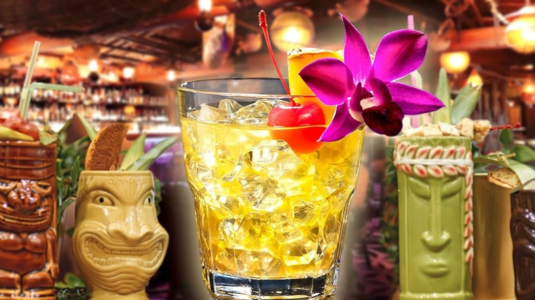 A Mai Tai garnished with a purple flower among several Tiki glasses