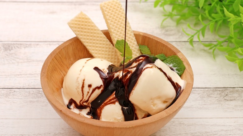 Chocolate sauce drizzling onto bowl of vanilla ice cream garnished with mint and wafers in wooden bowl.