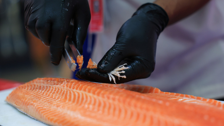 A person wearing black nitrile gloves pulls bones out of a salmon filet with sharp tongs