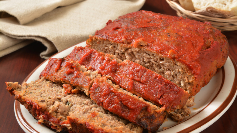 A partially sliced meatloaf with a thick red sauce on top, sitting on a white plate