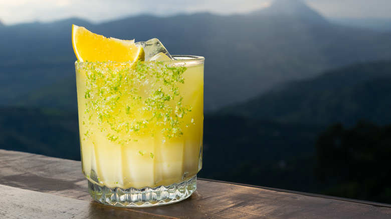 A yellow garnished cocktail served outside with mountains in the background.