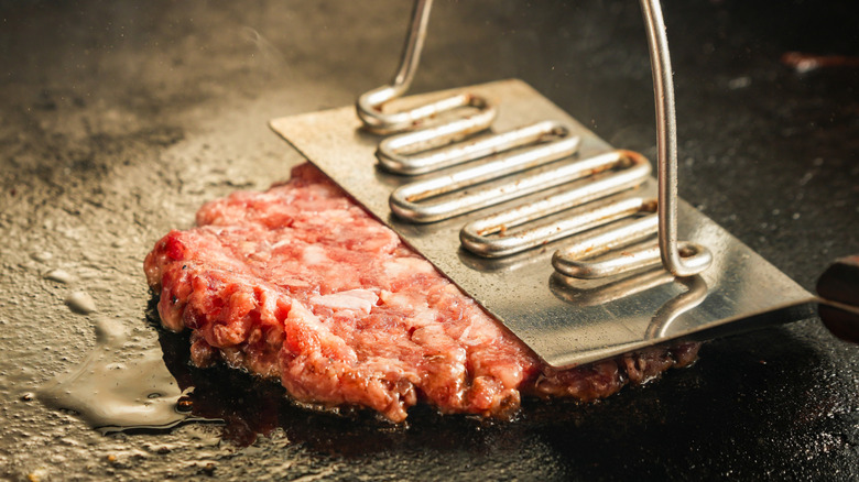 Raw beef patty being smashed on the grill with a metal utensil