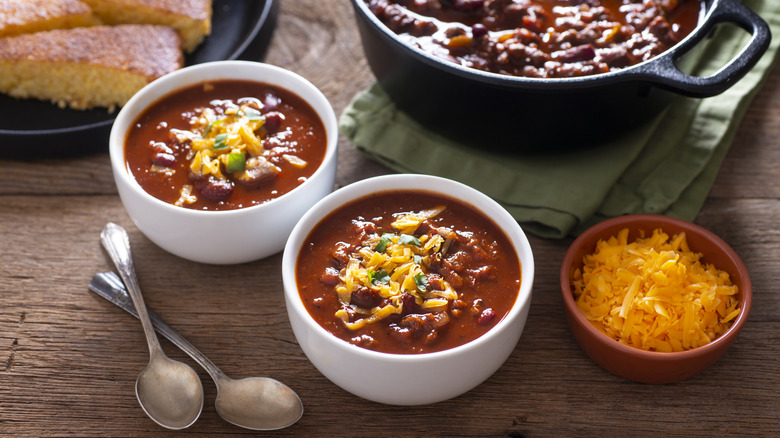 Two white bowls of chili with beans on a table with shredded cheese