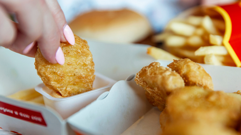 Fingers holding a Chicken McNugget dunking it in dipping sauce with more McNuggets and fries in the background