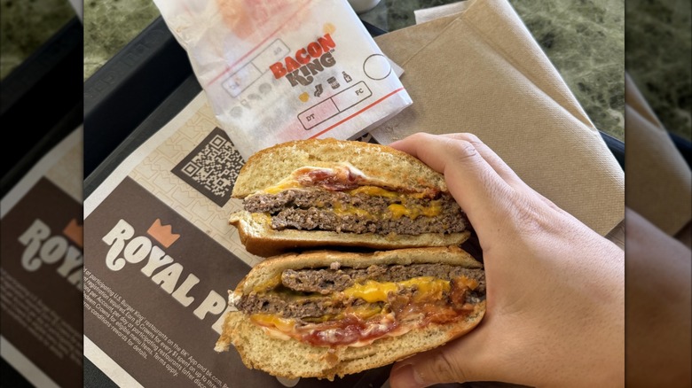 A person holding a Burger King Big King Burger that's been cut in half to show the cross section, over a paper lined tray, with the Bacon King of wrapper above