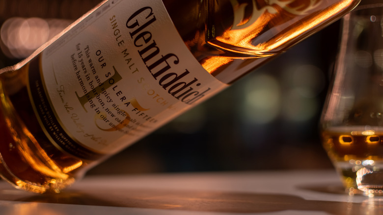 A tilted bottle of Glenfiddich 15 Year Single Malt Scotch with a blurry glencairn glass behind it.