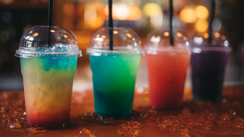 four colorful iced cocktails in plastic to-go cups on a bar with ice