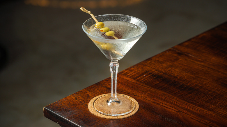 Cold dirty martini garnished with three olives sitting on a coaster on the corner of a wooden table