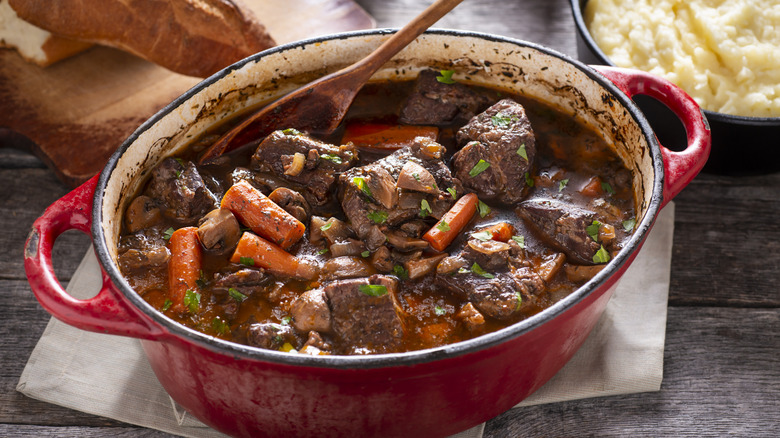 Beef stew in a red enameled cast iron Dutch oven, on a table with bread and mashed potatoes