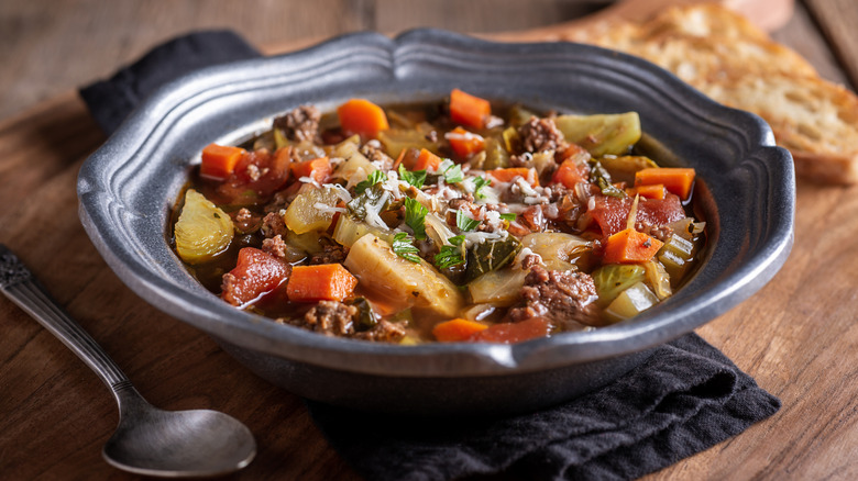 A blue bowl of hearty beef stew on a table next to a slice of toasted bread