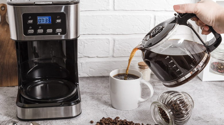 a hand pouring a cup of coffee from a the pot of a drip coffee maker beside the machine itself on a grey countertop