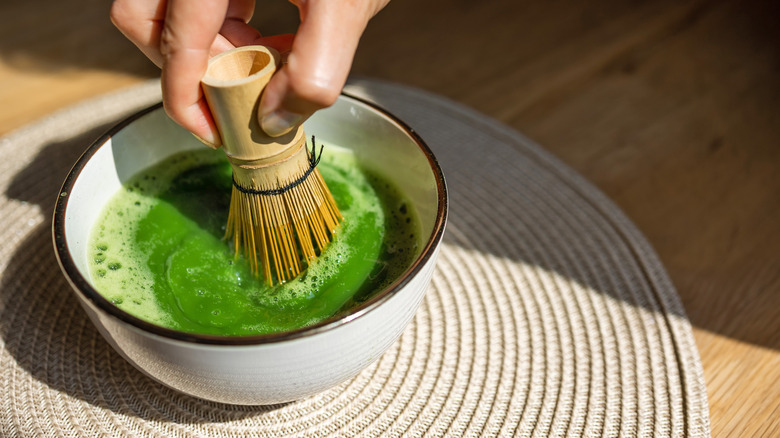 Someone whisking a bowl of bright green matcha with a bamboo whisk on a wooden table with a round woven table runner