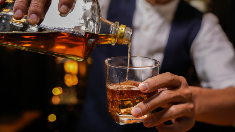 A bartender pours a glass of whiskey over ice from an unmarked bottle.