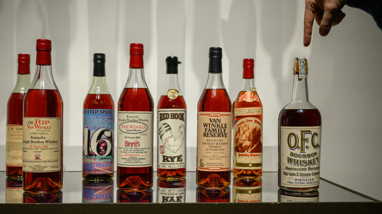 A line of bottles of bourbon on a glass table, with a finger pointing at the bottle on the far right