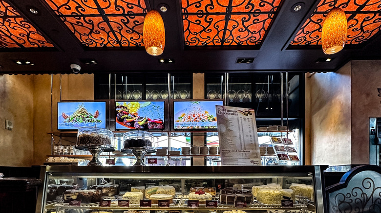 A Cheesecake Factory display case loaded with desserts underneath an orange glass ceiling with hanging orange lamps
