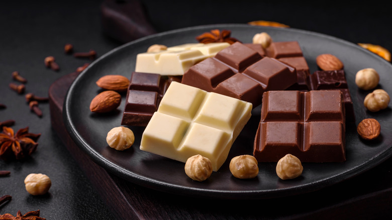 squares of different types of chocolate with nuts scattered beside them on a dark plate surrounded by whole spices