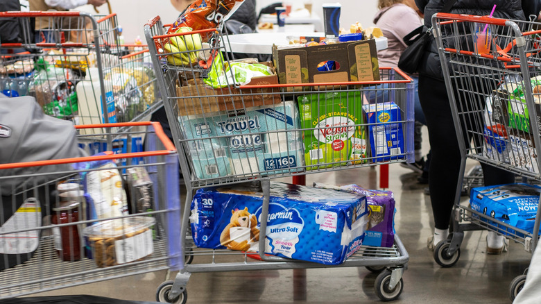 Shopping carts in line to check out at a Costco filled with items like paper towels, bananas, Doritos, and more
