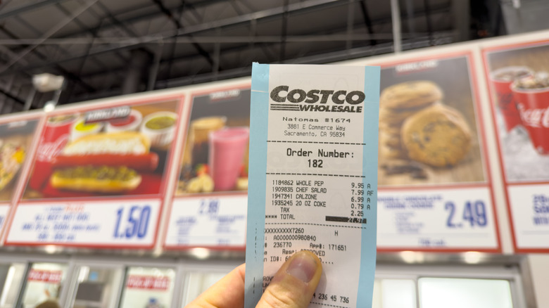 A person holding a Costco receipt showing several purchases in front of a Costco menu board