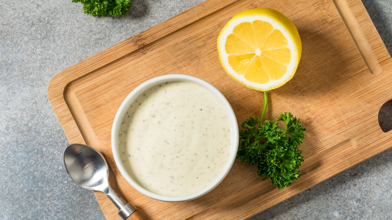 A bowl of ranch dressing sitting on a cutting board next to a sprig of parsley and half a lemon