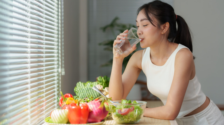 woman drinking water over a counter with fruits and veggies