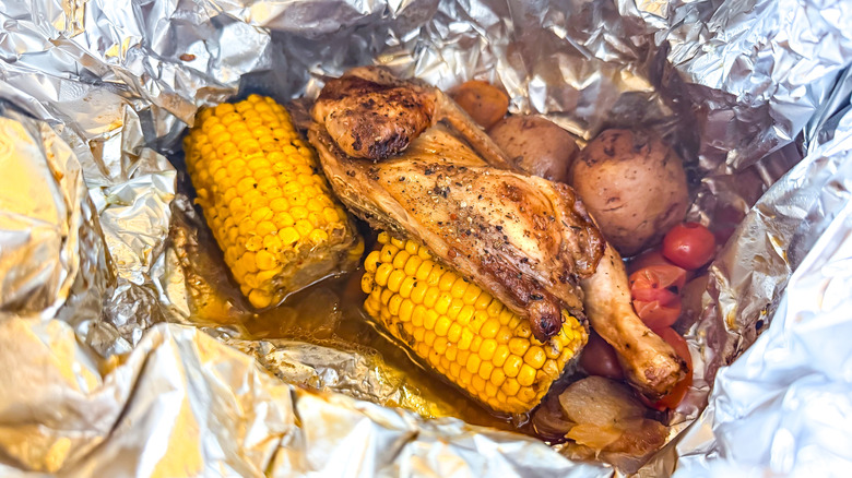 A meal cooked in aluminum foil with corn, chicken, potatoes, and small tomatoes