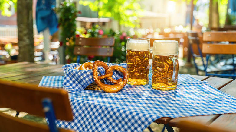 Two beers with frothy heads in dimpled mugs sit on a table at a German beer garden next to some German pretzels.