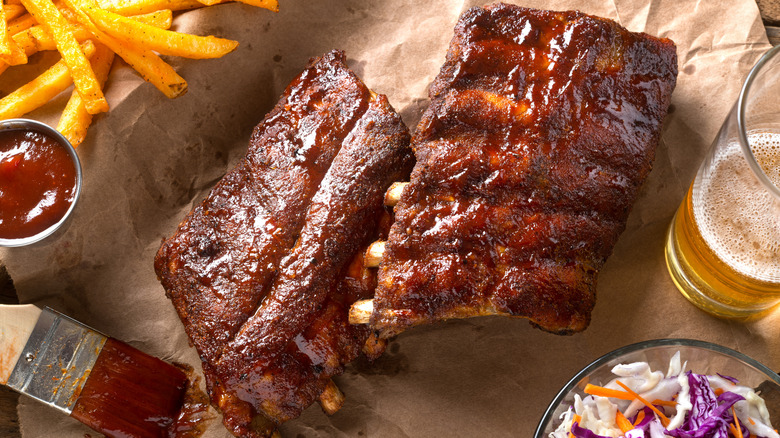 Two racks of BBQ ribs next to a glass of beer, French fries, a tub of sauce, and some coleslaw