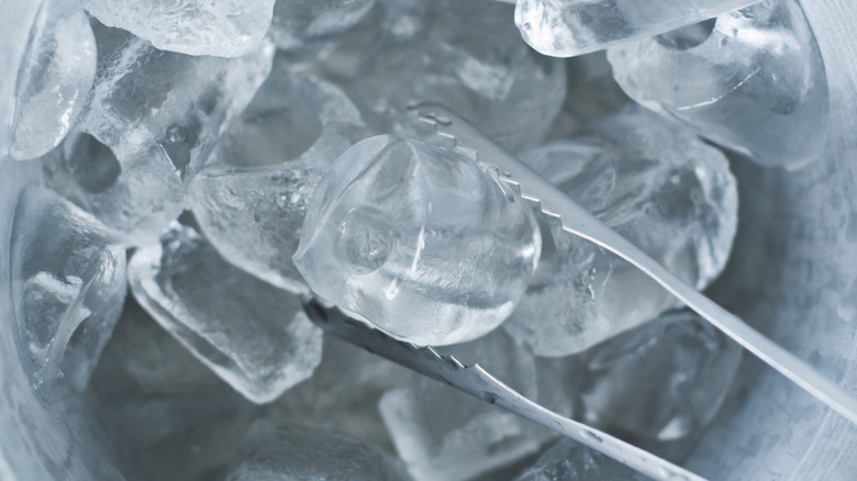 A bucket full of ice cubes with one grasped in a pair of metal tongs
